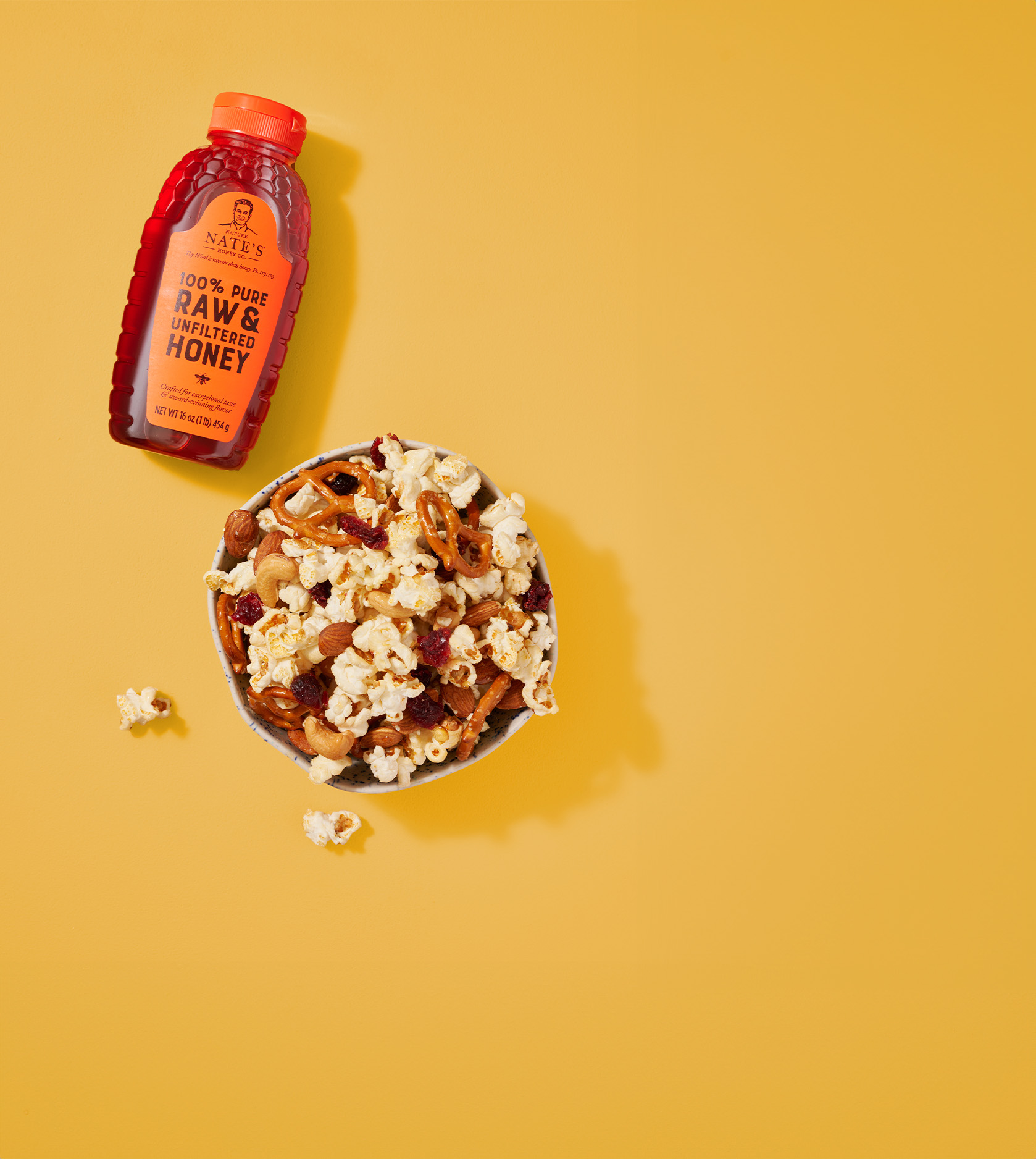 Nate's bottle of honey next to a bowl of popcorn, pretzels and more snack foods on a yellow background.