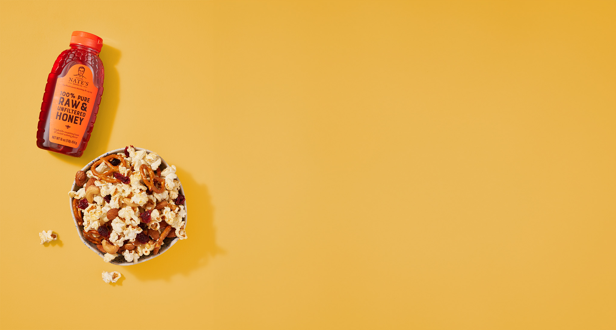 Nate's bottle of honey next to a bowl of popcorn, pretzels and more snack foods on a yellow background.