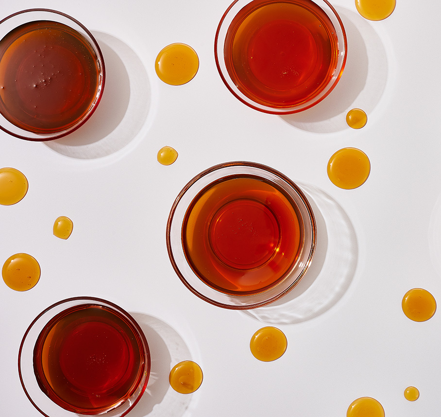 Overhead view of glass bowls filled with amber‑colored honey, with small drops of honey scattered on a white surface.